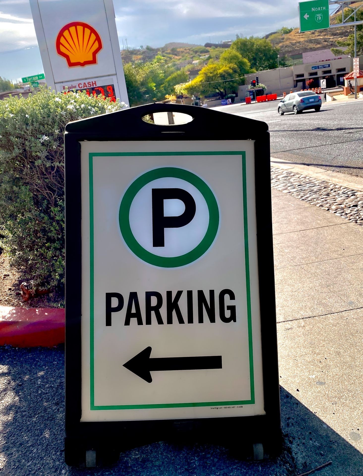 Outdoor parking sign with an arrow pointing left, Shell gas station and road in the background.